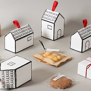 May include: Assortment of small gift boxes in the shape of houses with red ribbon accents. The boxes are white with black outlines and a simple design. Some boxes contain small treats like cookies and pastries.