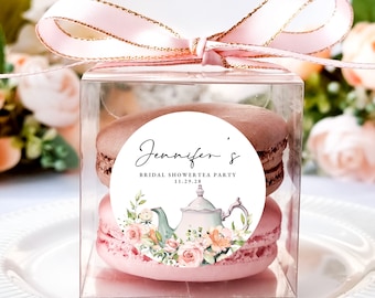 Puede incluir: Una caja de plástico transparente con una cinta y un lazo rosa, que contiene dos macarons rosas con una pegatina redonda blanca en la parte superior. La pegatina presenta una ilustración de acuarela de una tetera con flores y el texto "Jennifer's Bridal Shower Tea Party 11.29.28".