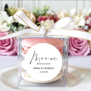 May include: A clear square box tied with a white ribbon with gold trim, containing two macarons. A white circular sticker on the box reads "Merci Beaucoup" with the names "Emma & Charles" and the date "11.24.29".