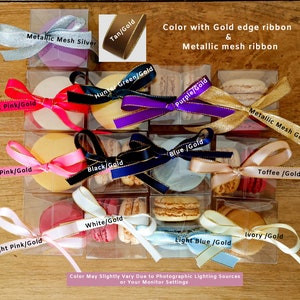 May include: Assortment of macarons in various colors, each with a gold-edged ribbon and a label indicating the color and gold combination. The labels include: Hot Pink/Gold, Metallic Mesh Silver, Tan/Gold, Hunter Green/Gold, Pink/Gold, Black/Gold, Purple/Gold, Blue/Gold, Metallic Mesh Gold, Toffee/Gold, White/Gold, Light Pink/Gold, Light Blue/Gold, and Ivory/Gold. The macarons are displayed in clear plastic boxes on a wooden surface.
