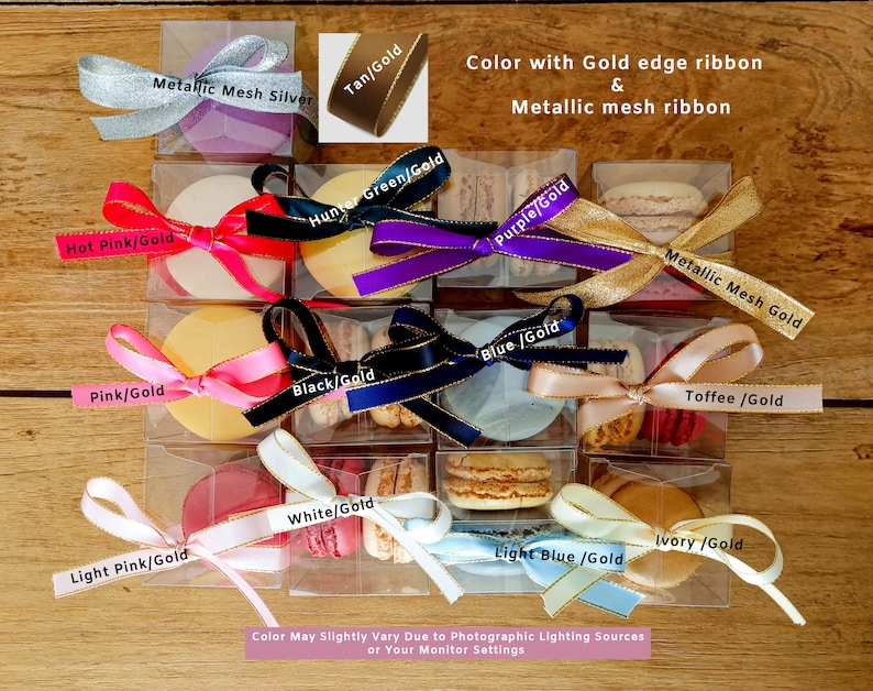 May include: Assortment of macarons in various colors, each with a gold-edged ribbon and a label indicating the color and gold combination. The labels include: Hot Pink/Gold, Metallic Mesh Silver, Tan/Gold, Hunter Green/Gold, Pink/Gold, Black/Gold, Purple/Gold, Blue/Gold, Metallic Mesh Gold, Toffee/Gold, White/Gold, Light Pink/Gold, Light Blue/Gold, and Ivory/Gold. The macarons are displayed in clear plastic boxes on a wooden surface.