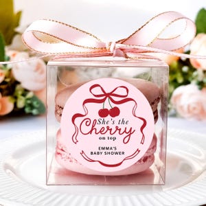 May include: A clear square box tied with a pink ribbon, containing two macarons. A pink circular sticker on the box reads "She's the cherry on top" with "Emma's Baby Shower" below. The macarons are pink and brown.