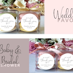 May include: Two clear boxes with macarons and a round sticker that says "a sweet thank you" with a heart. The boxes are tied with a gold ribbon and the text "Wedding Favor" is written in cursive above the boxes.