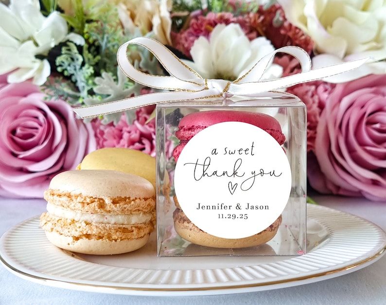 May include: A clear box filled with pastel-colored macarons, tied with a white ribbon with gold trim. A white round sticker with black text reads "a sweet thank you" with a heart symbol and the names "Jennifer & Jason" and the date "11.29.25".