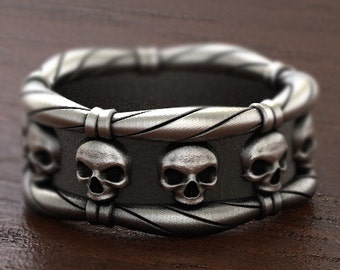 May include: A silver ring with a twisted band and five skull designs around the band.