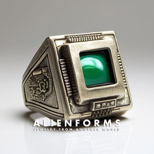 May include: A silver ring with a green gemstone set in a square frame. The ring has a detailed design with a textured surface and a raised border around the gemstone. The ring is a statement piece with a futuristic design.