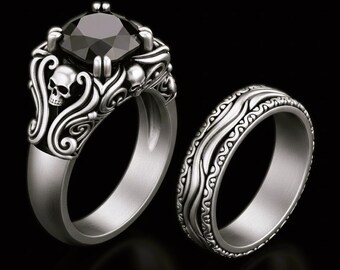 May include: A silver ring set featuring a large black gemstone and a matching band. The engagement ring has a skull design on the sides and intricate detailing. The band has a wavy pattern.