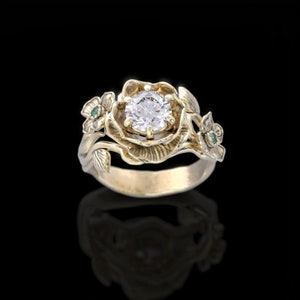 Op de afbeelding: Een gouden ring met een grote, ronde, heldere diamant gezet in een bloemmotief. De ring heeft twee kleinere groene edelstenen aan de zijkanten.