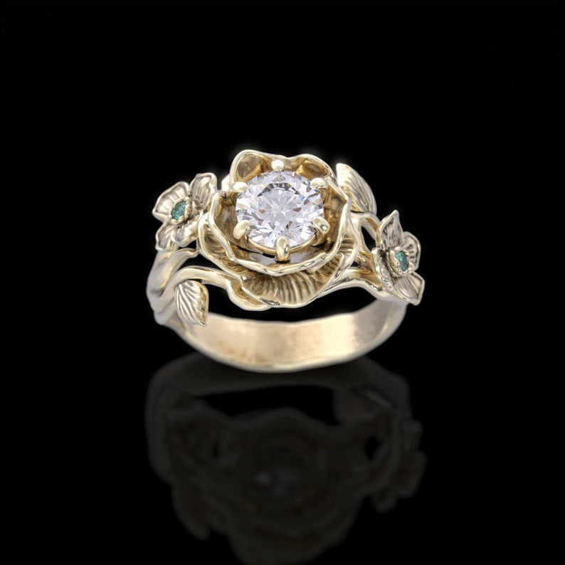 Op de afbeelding: Een gouden ring met een grote, ronde, heldere diamant gezet in een bloemmotief. De ring heeft twee kleinere groene edelstenen aan de zijkanten.