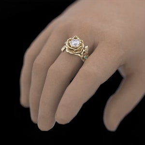 Op de afbeelding: Een gouden ring met een grote, heldere diamant gezet in een roosvormig ontwerp. De ring heeft ook groene edelstenen en delicate bladdetails.