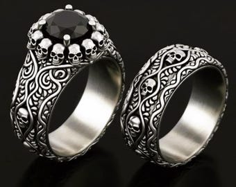 May include: Two silver rings with a gothic design. One ring features a large black gemstone surrounded by small skulls. The other ring has a band with a repeating skull and scrollwork pattern. The rings are set against a dark background.