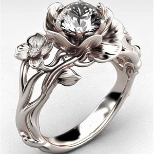 Op de afbeelding: Een zilveren ring met een grote, heldere edelsteen gezet in een bloemvormig ontwerp. De ring heeft ingewikkelde details en een delicaat bloemmotief.