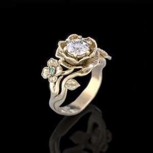 Op de afbeelding: Een gouden ring met een grote, heldere diamant gezet in een roosvormige setting. De ring heeft ook een kleinere groene edelsteen gezet in een kleinere bloem.
