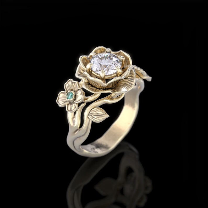 Op de afbeelding: Een gouden ring met een grote, heldere diamant gezet in een roosvormige setting. De ring heeft ook een kleinere groene edelsteen gezet in een kleinere bloem.