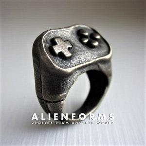 May include: A silver-toned ring shaped like a video game controller. The ring has a textured surface and features a cross-shaped button and two round buttons.