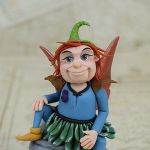May include: A whimsical fairy figurine with red hair, a green hat, and blue wings, sitting on a grey rock. The fairy is wearing a blue dress with a green skirt and yellow shoes.