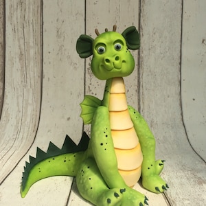 May include: A green fondant dragon figurine with yellow stripes, black eyes, and a small wing on its back. The dragon is sitting on a white wooden surface.
