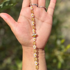 Könnte beinhalten: Ein Perlenarmband mit abwechselnden weißen und gelben Gänseblümchen-Perlen sowie durchscheinenden bernsteinfarbenen facettierten Perlen. Ein kleiner goldfarbener Metall-Bienenanhänger mit schwarz-weißen Emaille-Details befindet sich in der Mitte.