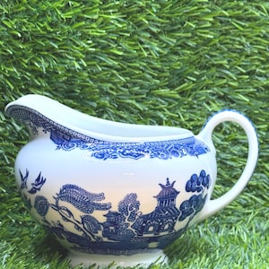 May include: White ceramic creamer with a blue Willow pattern design. The creamer features a spout, a handle, and a detailed landscape scene with pagodas, trees, and birds. The rim and handle are accented with blue patterns. The creamer sits on a small foot.