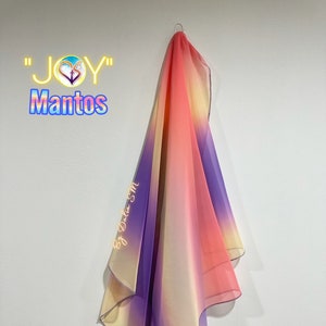 May include: A sheer, colorful scarf with a gradient of colors, including pink, orange, yellow, and purple, hangs against a white wall. The scarf is labeled "JOY Mantos" with text that reads "By Dalia SM".