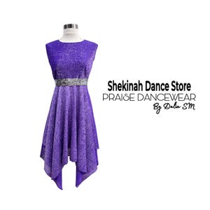 May include: A sleeveless, purple dance dress with a shimmering fabric. The dress features a silver belt and a unique asymmetrical hemline. The text "Shekinah Dance Store PRAISE DANCEWEAR By Dalia SM" is visible on the right side.