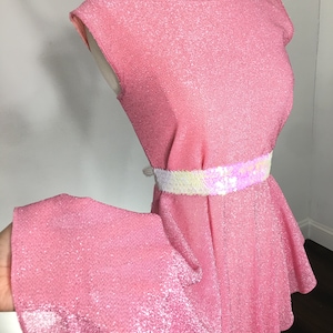 May include: A pink sparkly dress with a white and iridescent sequin belt. The dress has a ruffled skirt and a sleeveless bodice.