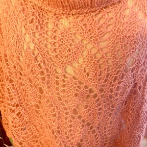 May include: A peach-coloured knit garment featuring an openwork lace pattern with a prominent cowl neckline. The fabric displays a delicate, airy texture with intricate diamond-shaped motifs throughout the weave, creating a semi-sheer appearance.