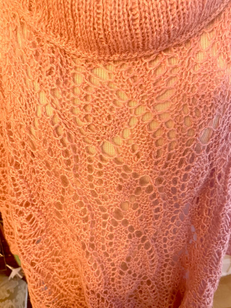 May include: A peach-coloured knit garment featuring an openwork lace pattern with a prominent cowl neckline. The fabric displays a delicate, airy texture with intricate diamond-shaped motifs throughout the weave, creating a semi-sheer appearance.