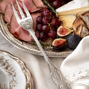 May include: A silver-toned metal serving fork with an ornate handle featuring embossed shell and leaf motifs. The fork rests on a silver platter filled with sliced roast beef, red grapes, wedges of brie cheese, and halved fresh figs.