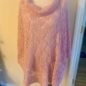 May include: A light pink knit poncho featuring a prominent cowl neckline and an intricate openwork lace pattern throughout the body. The garment has a relaxed, draped silhouette with ribbed edges at the collar and hem, showcasing a delicate texture.