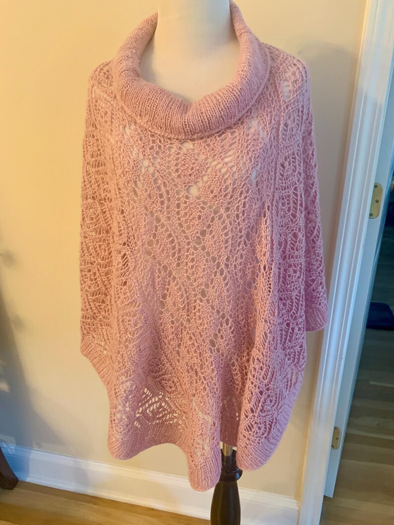 May include: A light pink knit poncho featuring a prominent cowl neckline and an intricate openwork lace pattern throughout the body. The garment has a relaxed, draped silhouette with ribbed edges at the collar and hem, showcasing a delicate texture.