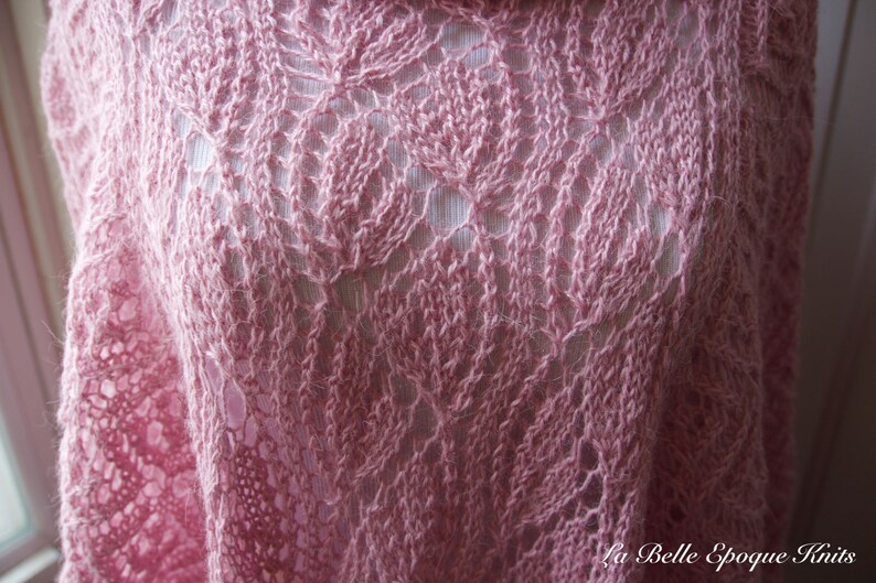 May include: A soft pink knitted shawl featuring an intricate openwork leaf pattern throughout the fabric. The garment displays a delicate, airy texture with visible yarn loops and a repeating botanical motif. The text La Belle Epoque Knits is visible.