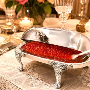 May include: A silver-plated serving dish with a domed lid, featuring ornate lion-head feet and intricate floral detailing. The dish contains a glass insert filled with bright red caviar and includes a small white serving spoon resting inside.