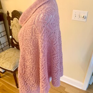 May include: A light pink knitted poncho featuring a prominent cowl neckline and an intricate openwork lace pattern throughout the body. The garment is constructed with a ribbed hem and collar, showcasing a delicate texture and soft yarn finish.