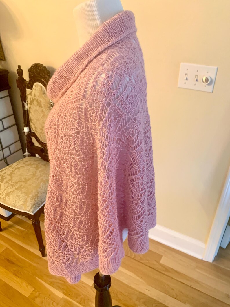 May include: A light pink knitted poncho featuring a prominent cowl neckline and an intricate openwork lace pattern throughout the body. The garment is constructed with a ribbed hem and collar, showcasing a delicate texture and soft yarn finish.