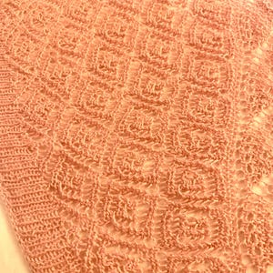 May include: A peach-coloured knitted textile featuring a repeating square pattern with an openwork design. The fabric displays a textured, grid-like structure created by interconnected loops, showcasing a consistent geometric motif throughout the piece.