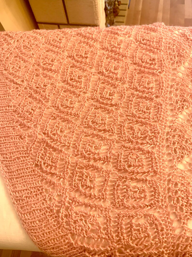 May include: A peach-coloured knitted textile featuring a repeating square pattern with an openwork design. The fabric displays a textured, grid-like structure created by interconnected loops, showcasing a consistent geometric motif throughout the piece.