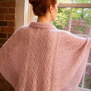 May include: A light pink knitted poncho featuring an intricate lace pattern with diamond and vertical motifs. The garment has a ribbed collar and a wide, flowing silhouette that drapes over the shoulders, showcasing a delicate, open-work texture.