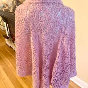 May include: A light pink knitted poncho featuring an intricate openwork lace pattern throughout the body. The garment has a solid knit collar and a matching solid knit hemline, providing a structured finish to the delicate, airy texture.