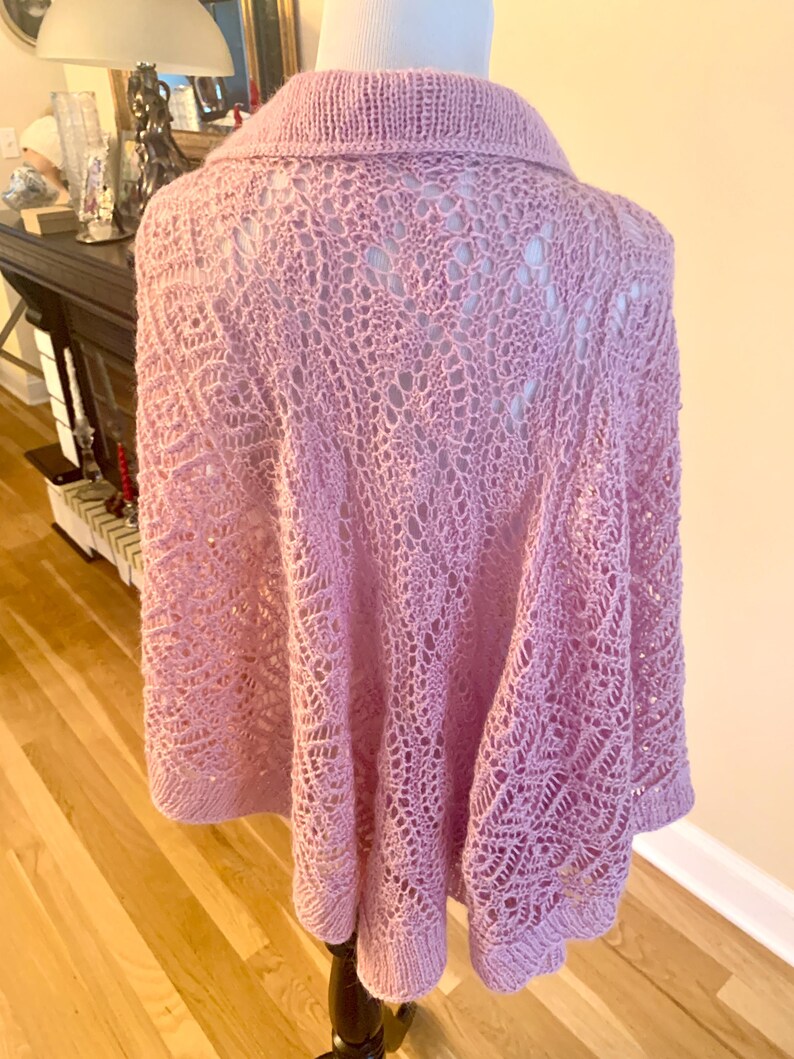 May include: A light pink knitted poncho featuring an intricate openwork lace pattern throughout the body. The garment has a solid knit collar and a matching solid knit hemline, providing a structured finish to the delicate, airy texture.