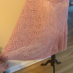 May include: A light pink knit poncho featuring a cowl neckline and an intricate openwork pattern. The garment displays a combination of square-shaped motifs and delicate lace-like textures throughout the fabric, creating a soft and airy appearance.