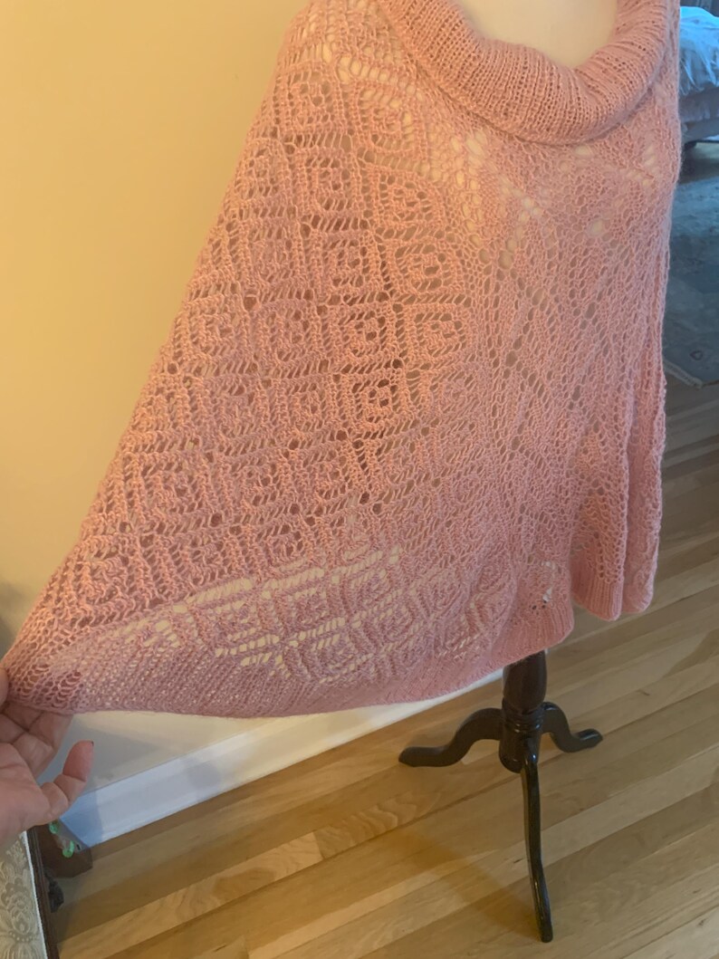 May include: A light pink knit poncho featuring a cowl neckline and an intricate openwork pattern. The garment displays a combination of square-shaped motifs and delicate lace-like textures throughout the fabric, creating a soft and airy appearance.