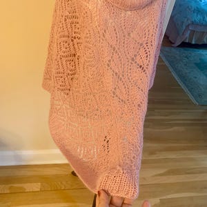 May include: A light pink knit poncho featuring a cowl neckline and an intricate openwork lace pattern throughout the body. The garment has a ribbed hem and a loose, draped silhouette that hangs from the shoulders, showcasing a delicate yarn texture.
