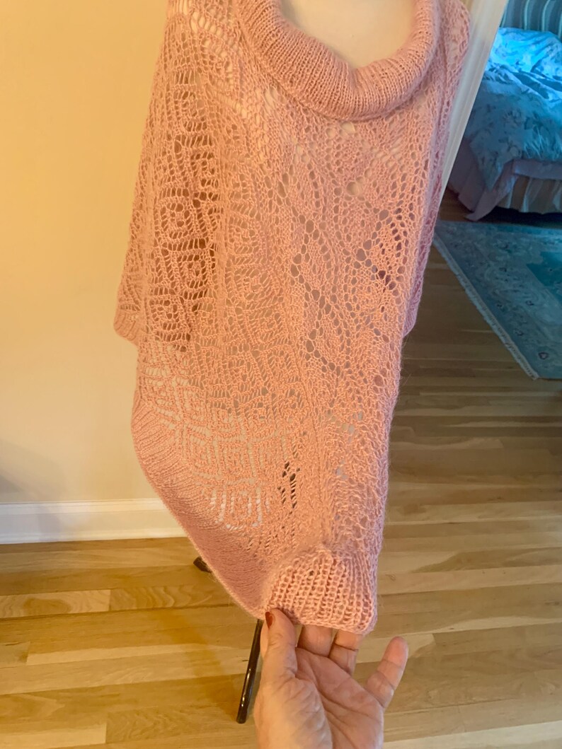 May include: A light pink knit poncho featuring a cowl neckline and an intricate openwork lace pattern throughout the body. The garment has a ribbed hem and a loose, draped silhouette that hangs from the shoulders, showcasing a delicate yarn texture.