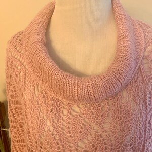 May include: A light pink knit poncho featuring a wide, ribbed cowl neckline and an intricate, openwork lace pattern throughout the body. The garment is crafted from soft yarn, showcasing a delicate, airy texture with a consistent pastel hue.