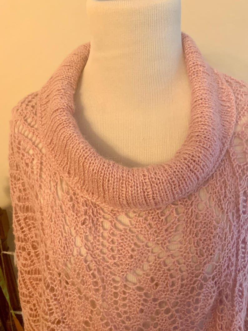 May include: A light pink knit poncho featuring a wide, ribbed cowl neckline and an intricate, openwork lace pattern throughout the body. The garment is crafted from soft yarn, showcasing a delicate, airy texture with a consistent pastel hue.