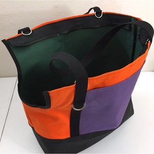 May include: A large tote bag with a color-block design. The bag features orange, purple, and black panels, with a black handle and a green interior. The bag has a zippered top and metal rings for the handles.