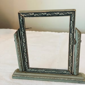 Pode incluir: Uma moldura de foto vintage azul claro com uma abertura retangular. A moldura tem um design ornamental e é suportada por dois painéis laterais e uma base. A moldura é articulada e ajustável. A moldura é de metal.