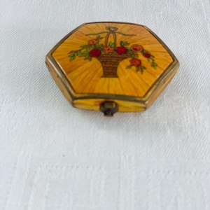 Puede incluir: Compacto hexagonal vintage con un diseño de cesta floral. El compacto presenta un fondo amarillo con un patrón radiante, un marco color latón y un cierre. La cesta floral contiene flores rojas y naranjas.