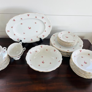 Könnte beinhalten: Ein weißes Porzellan-Tafelservice mit zartem Blumenmuster. Das Set umfasst eine ovale Servierplatte, Teller, Schalen, Teetassen und Untertassen. Jedes Stück hat einen goldenen Rand und kleine rosa Rosenakzente.